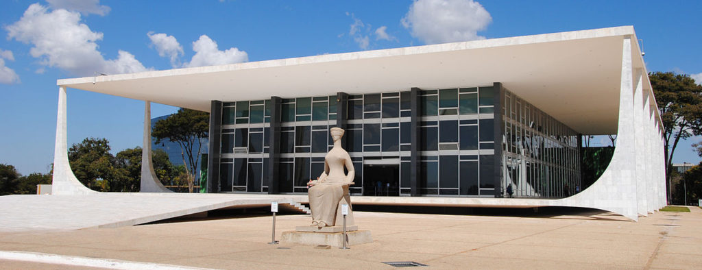 Sede do Supremo Tribunal Federal, edifício suspenso próximo ao chão com colunas modernas brancas e caixa de vidro escuro, com estátua da justiça sentada à frente.